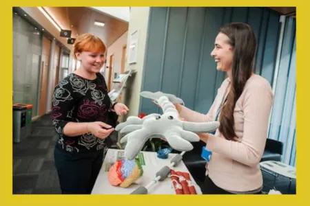 two women smiling at each other, one holding soft toy of a brain cell (neuron)